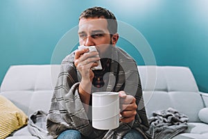 Sick young man blowing her nose using facial tissues and holding white mug