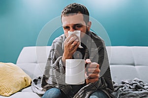 Sick young man blowing her nose using facial tissues and holding white mug
