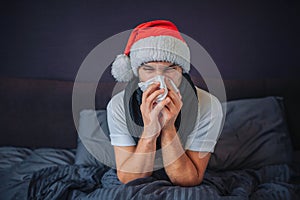 Sick young man in christmas red hat sits on bed. He is covered with blanket. Guy sneezing into tissue. He suffers. Young