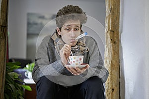 A sick young man with a cold sitting in his house has a thermometer in his mouth and is holding a cup in his hand