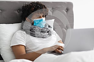 Sick young man in face mask staying in bed, using laptop