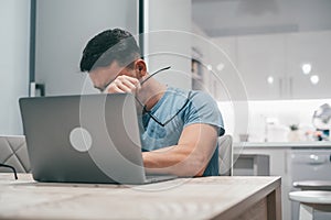 Sick young man feeling unhealthy at home working with laptop on the table alone. Teenager boy taking off his glasses after tired