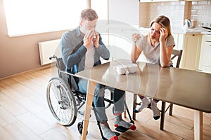 Sick young man with inclusiveness sneezing with healthy woman at table. Sick people in kitchen. Headache and pain