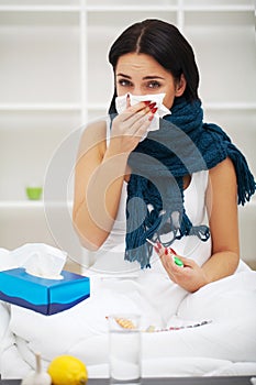 Sick young woman at home on the sofa, she is covering with a blanket, taking temperature and blowing her nose with a tissue