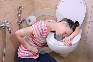 Sick young woman with pain in stomach is vomiting in toilet sitting on the floor at home, food poisoning symptom.