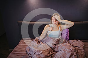 Sick young woman sits on bed and holds one hand on forehead. She holds glass of water and looks at it. Model is suffer