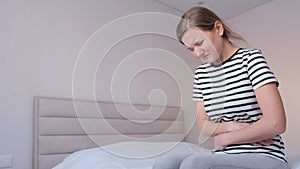 A sick young woman sits on the bed at home, touches her stomach, suffers from abdominal pain.