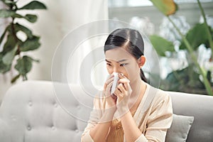 Sick young woman sitting on sofa blowing her nose at home in the sitting room. Photo of sneezing woman in paper tissue