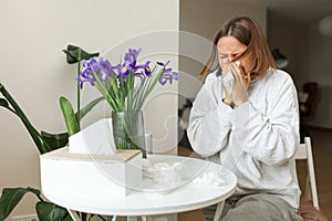 Sick young woman suffering from allergy, fever or flu blows nose and sneezing in paper tissues, sits by table at home