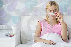 Sick young woman taking temperature with thermometer in bed