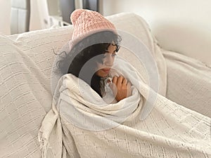Sick young woman tucked in a blanket on her sofa, very cold due to the low temperature in her home on a winter day. Cold concept