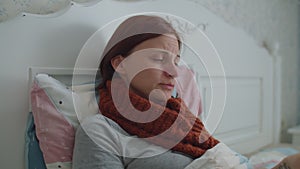 Sick young woman with warm scarf on neck laying in bed coughing and measuring her temperature with electronic infrared