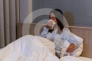 Sickness Asian woman sneezing into a tissue in bed room at night. Female illness from colds or flu. Allergy or health issue