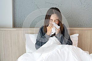 Sickness Asian woman sneezing into a tissue in bed room after wake up. Female illness from colds or flu. Allergy or health problem