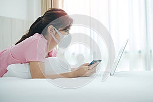 Sickness woman with face mask holding credit card and using computer laptop for online shopping on bed in morning at home. New