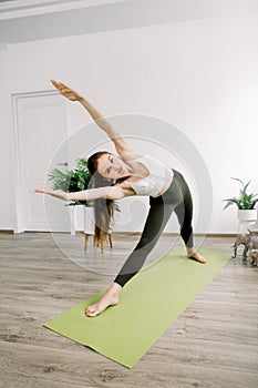 Side angle full length view of attractive young sporty woman, practicing sport and yoga, standing on green yoga mat in
