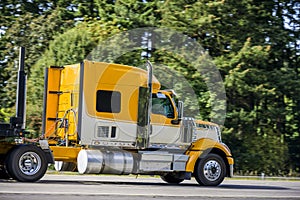 Side of big rig yellow semi truck with semi trailer running on the flat road with green trees on the shoulder