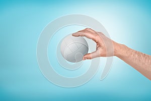 Side close-up of man`s hand holding a little white volleyball on light-blue background.