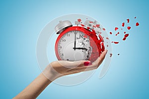 Side closeup of woman`s hand facing up and holding red retro alarm clock that is dissolving into small pieces on light