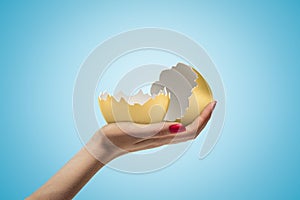 Side closeup of woman`s hand facing up and holding two parts of empty gold eggshell on light blue gradient background.