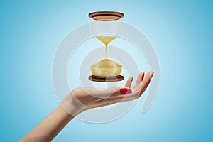 Side closeup of woman`s hand facing up and levitating hourglass on light blue background.