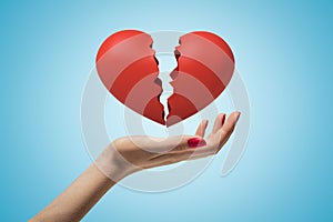 Side closeup of woman`s hand facing up and levitating red broken heart on light blue gradient background.