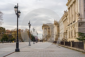 Side facade of the royal palace of Madrid, pedestrian street with lampposts, trees and sunny day with clouds