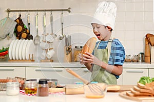 Side of face kid eating large loaves of bread, happy Asian boy with apron and chef hat holding and eating freshly baked homemade