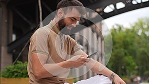 Side low-angle view of cheerful young man sitting on bench outdoors using typing smartphone. Confident bearded male