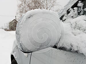 The side mirror of the car is covered with snow during a gloomy winter day