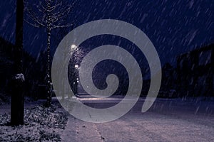 A side night portrait of a snowy street during heavy snowfall with street lights illuminating the way for cars. The road looks