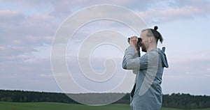 Side portrait of Caucasian man with bun holding binoculars outdoors green field background. Bearded tourist looking