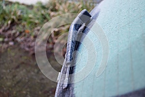 A side portrait of a frozen back window of a car with a windshield wiper frozen stuck during winter. The ice first needs to melt