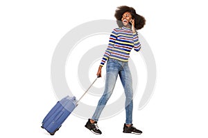 Side portrait of happy young black woman walking with cellphone and suitcase against isolated white background