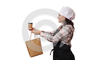 Side portrait, nice waitress in apron and chef hat, smiles holding out hot takeaway drink in cardboard cup and paper bag