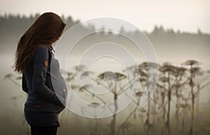 Side portrait of pregnant woman with long hair. Woman with big belly looking down. Foggy misty evening landscape
