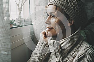 Side portrait of serene female looking outside the window at home in winter season Cold house temperature. One woman wearing knit