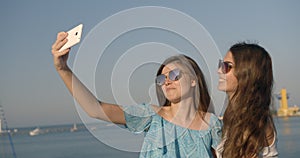 Side portrait of two young adorable girlfriends making funny faces while taking selfies via the mobile phone on the