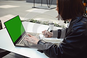 Side portrait of a woman freelancer, copywriter, writing texts on notepad, typing on keyboard of a laptop computer with a green