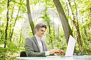 Side Portrait of young handsome business man in suit working at laptop at office table in green forest park. Business concept.