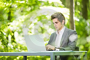 Side Portrait of young handsome business man in suit working at laptop at office table in green forest park. Business concept.