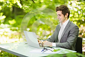 Side Portrait of young handsome business man in suit working at laptop at office table in green forest park. Business concept.