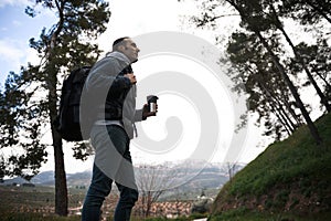 Side portrait of a young man with backpack and travel mug, during a trekking in the forest, mountains on the background