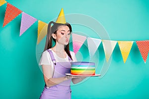 Side profile photo portrait of happy birthday girl wearing cone keeping cake with pouted lips isolated on vivid teal
