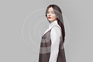 Side profile view portrait of serious beautiful young woman in white shirt and brown apron with makeup and brunette hair standing