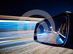 In the side rear-view mirror of the car, the evening road and lights are reflected