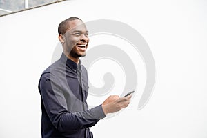 Side of smiling African American man holding mobile phone by white wall