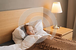 Side view of adorable little sick girl lying in bed measuring temperature with thermometer in mouth at home.