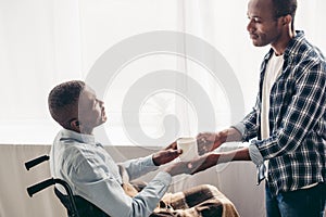 side view of adult son giving cup of hot beverage to disabled senior father photo