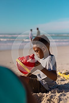 Side view of african american boy holding pail playing with sand on beach against sea and blue sky